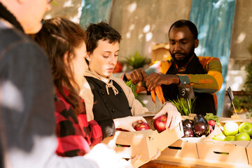 Team of multicultural merchants selling fresh fruits and veggies in boxes, serving elderly man and young woman at local farmers market. Diverse people looking at healthy organic colorful produce.