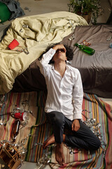 High angle of sick man with hangover symptoms and intoxication keeping head on edge of bed while sitting on the floor
