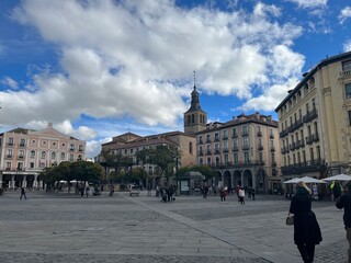 Obraz premium Scenic European town square with historic buildings, church tower, outdoor cafés, and pedestrians enjoying a bright day under a partly cloudy sky