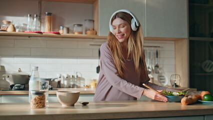 Earphones girl mixing salad listening music at kitchen. Lady preparing breakfast