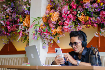handsome male businessman with glasses wearing headphones annoyed operating cellphone operating cellphone in front of laptop computer on table, sitting in indoor cafe. for technology, lifestyle