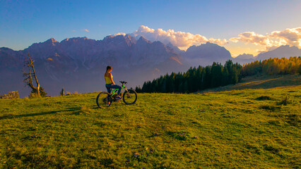 AERIAL: Young Caucasian woman mountain biking in the beautiful Julian Alps stops to marvel at the breathtaking golden sunset. Female tourist riding her electric bicycle rests and takes in the sights.