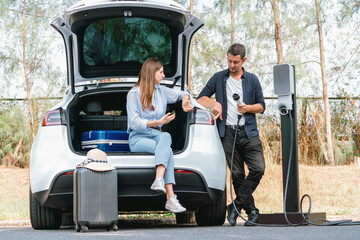 Lovely young couple recharging battery for electric car while relaxing and sitting on the trunk during road trip travel EV car in autumnal forest. Eco friendly travel on vacation during autumn. Exalt