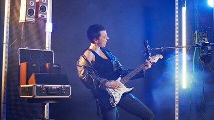 Obraz premium Rebellious edgy woman playing guitar in a modern studio setting, performing an iconic and bold song. Independent energetic artist singing with confidence at microphone, music lover. Camera A.