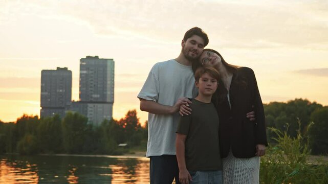 Portrait of a happy family of three, consisting of a mother, father, and child, is shown standing in a city park. In background, a newly constructed high-rise apartment building is visible, in which f