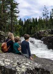 A mother and her two sons sit on a rock, captivated by the powerful Sognstupet Waterfall in Idre Dalarna surrounded by green trees and rushing water in the wilderness of Sweden