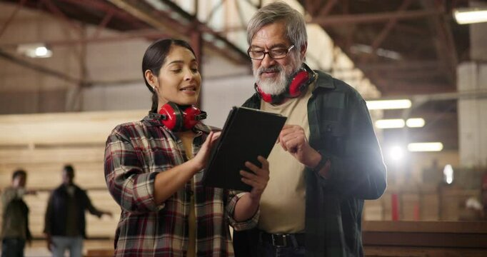 Carpentry, tablet and woman with mature mentor at factory for construction, help or building design advice. Wood, warehouse and apprentice consulting artisan expert with digital, plan or checklist