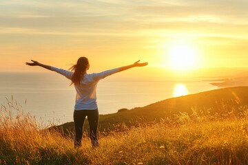 Woman with arms outstretched on a grassy hill, basking in the warm golden light of a beautiful sunrise over the ocean, embodying happiness and peace.