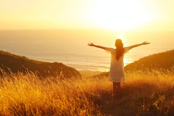 Woman with arms outstretched on a grassy hill, basking in the warm golden light of a beautiful sunrise over the ocean, embodying happiness and peace.
