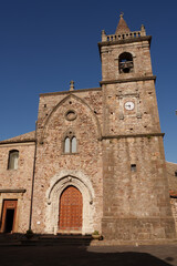 A historic Gothic-style stone church with a prominent bell tower in a Mediterranean village, showcasing intricate architecture and ancient cultural heritage in a serene setting