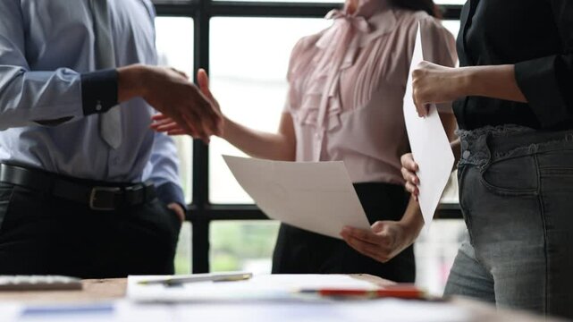 Business professionals discussing documents and shaking hands in office, symbolizing teamwork and collaboration during a meeting.