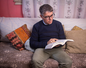 Adult man wearing glasses, entertaining himself by reading, sitting on the armchair