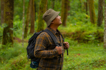 Half-body close-up of an adventurer in a dense and green pine forest. An Asian male traveler in a flannel shirt, bucket hat and backpack poses against a backdrop of dense pine trees.