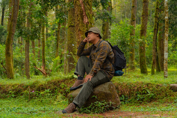 An adventurer is sitting on a large rock in a dense pine forest. An Asian man in a flannel shirt and bucket hat is sitting and resting while enjoying the tranquility of the beautiful nature.