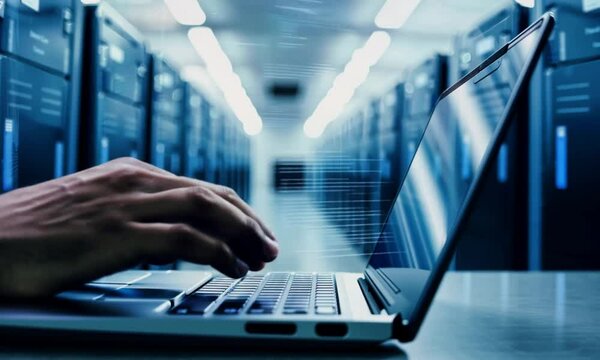 Close-up of hands typing on a laptop keyboard in front of a server rack in a data center.