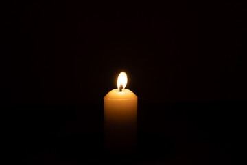 A single burning candle flame or light glowing on a big white candle isolated on red or dark background on table in church for Christmas, funeral or memorial service