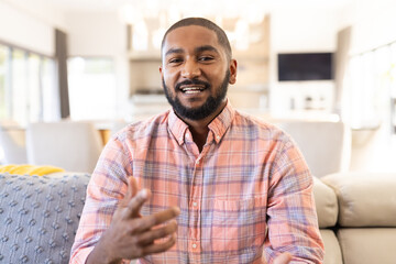 Talking and gesturing, smiling indian man in casual shirt sitting on couch, on video call