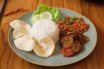 a plate of food with rice and stir-fried lung on top. with a complement of crackers, lettuce and cucumber. food on a brown wooden table