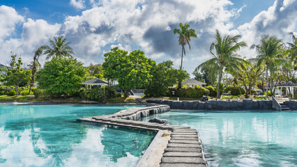 A winding concrete footpath crosses the swimming pool with clear turquoise water. In the distance, on the shore there is green tropical vegetation, palm trees, gazebos. Blue sky, clouds. Philippines.