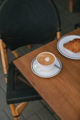 A delicious cup of cappuccino with a freshly baked croissant on a plate on the table in the cafe's outdoor terrace.