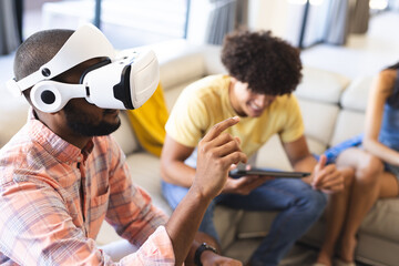 Using VR headset, man interacting with virtual environment while friend using tablet, at home