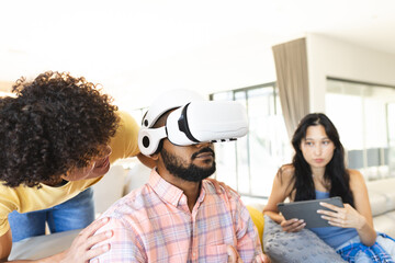 Using VR headset, man interacting with diverse friends, woman holding tablet nearby, at home