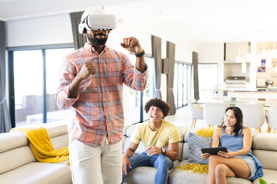 Using VR headset, man playing virtual game while diverse friends watching and laughing, at home