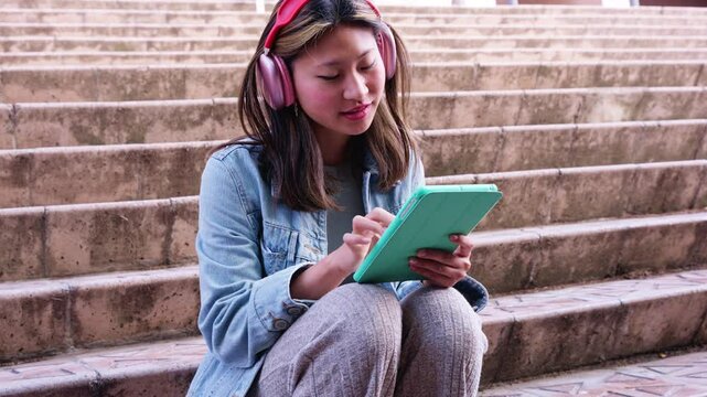 young chinese girl with headphones,sitting on university campus using social media app on digital tablet with surprised face