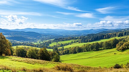 Fototapeta premium A panoramic view of rolling green hills and forested mountains under a bright blue sky with fluffy white clouds.