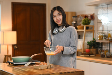 Smiling young asian woman making scrambled eggs for breakfast in cozy kitchen