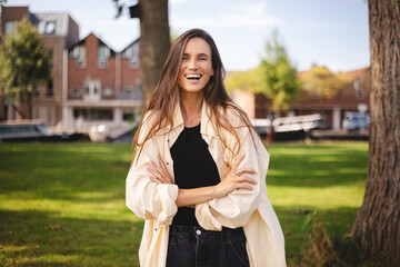 Smiling beautiful brown haired young woman wearing shirt and top 
standing outdoor in park posing...