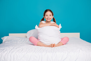 Full length photo of adorable little girl hugging pillow sit bed sleepover bedroom dressed stylish pajama isolated on blue color background