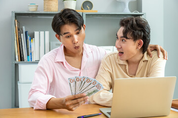 Two Asian men gay couple surprised by financial success while holding cash banknote and using laptop