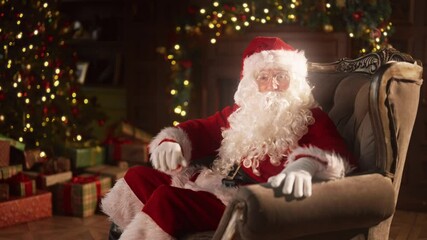 Santa Claus telling fairytales, congratulations and greeting, portrait in home. Old man in Santa costume talking to camera, sitting alone in armchair in cozy decorated room in Xmas eve, slow motion