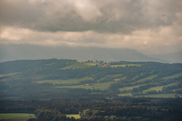 Fototapeta premium A misty, atmospheric view of rolling hills, forests, and scattered farms in Bavaria, with dark clouds covering the sky..