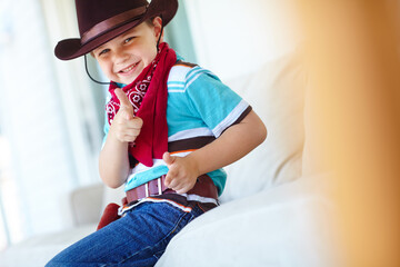 Kid, boy and portrait with cowboy costume in home for fantasy play, shooting game or pointing pretend gun. Person, smile or hand gesture on sofa for halloween, dress up or protection with imagination