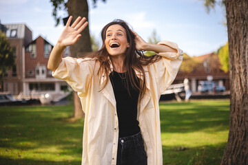 Brunette woman with long hair congratulating her friend, jumping, running to meet. Carefree friendly woman, smiling broadly while waving raised palm, greeting friend, meeting with mates walking park © zvkate