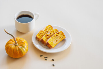Slices of pumpkin sweet bread or cake with icing on a plate on a light background with decorative pumpkin and a cup of coffee.