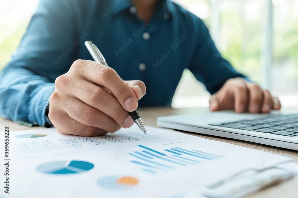 Poster close-up of a person writing notes while analyzing business graphs and charts on a desk with a lapto
