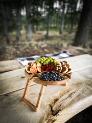 A wooden plate with fruits and cheeses on the background of a field with sheep. The concept of a picnic in nature