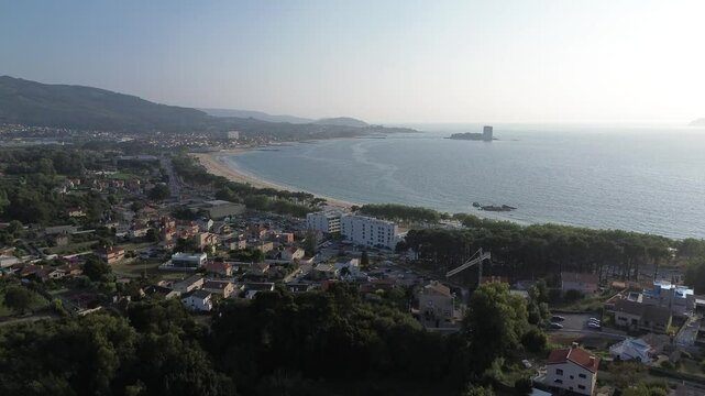Camera ascends revealing Samil Beach, bordered by a line of trees with buildings behind. The calm sea extends towards the horizon with Toralla Island in the distance on a sunny summer afternoon.