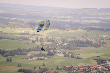 A paraglider soars above the green fields and forests of Bavaria, with a scenic view of the rural...