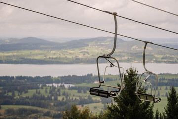 A close-up of an empty ski lift chair suspended over a scenic view of the Bavarian countryside,...