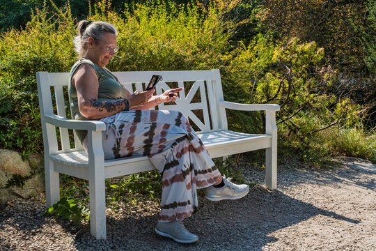 Female tattooed best ager enjoys a sunny autumn day in the park on a bench and takes photos with her smartphone