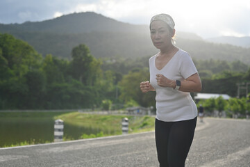 A focused senior woman jogs on a scenic path, surrounded by nature, with mountains in the distance, embodying strength, dedication, and the joy of staying active