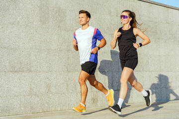 Young Man and Woman Jogging Together on a Sunny Day Near a Textured Concrete Wall in an Urban Setting