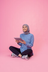 A cheerful woman in a colorful hijab enjoys her tablet while seated against a pink backdrop