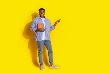 Fototapeta premium Full length photo of funky cool guy dressed checkered shirt holding book pointing emtpy space isolated yellow color background