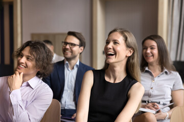 Fototapeta premium Joyful group of employees having fun at conference meeting, sitting in lines, looking at speaker away, listening to presenter. Business team attending career training, educational seminar