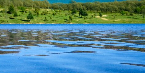 Rippling Waterscape, Gentle ripples on a calm blue lake reflect the surrounding lush green hills and trees. Selective focus.
